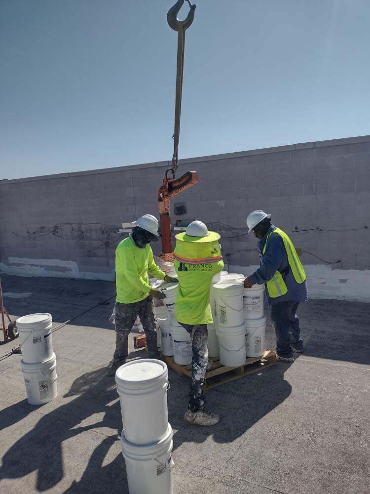 Two contractors are unloading buckets of white roof coating that have been delivered to the roof by a crane
